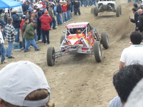 1990 Ford Bronco - red off-road buggy lifts its front wheels off a dirt rise during a desert race, with another car and a crowd behind it.