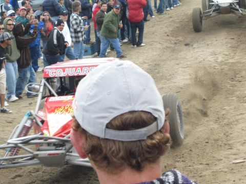 1990 Ford Bronco - rear of a red, flame-painted off-road buggy kicks up dust on a dirt course, viewed past a spectator in a backward white cap.
