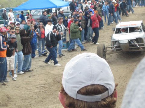 1990 Ford Bronco - white off-road race truck approaches on a dirt desert course as a large crowd watches from behind track markers.