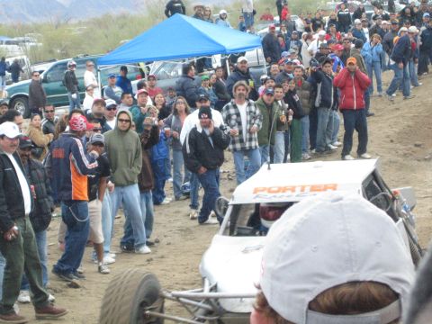 1990 Ford Bronco - white off-road race vehicle lettered 'Porter Racing' sits on a dirt course while bundled-up spectators look on under a blue canopy.