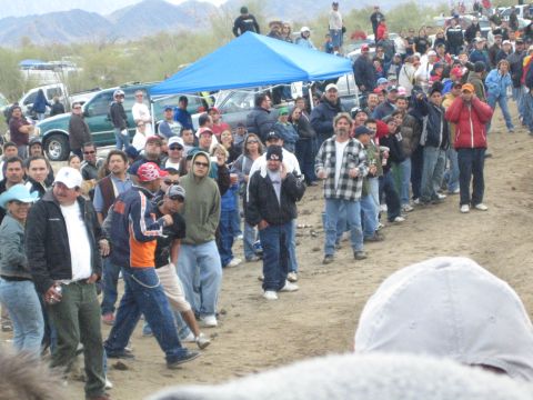 1990 Ford Bronco - Spectators in winter jackets gather along a desert race course near a blue pop-up canopy, with mountains in the distance.