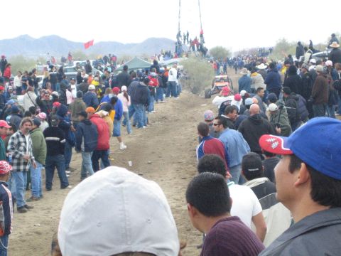 1990 Ford Bronco - wide view down a dirt desert race course lined on both sides by large crowds of spectators under overcast skies.