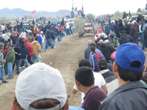 1990 Ford Bronco - orange off-road buggy drives a dirt desert course in the distance, flanked by long rows of spectators wearing caps and jackets.