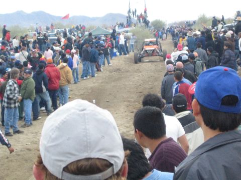1990 Ford Bronco - off-road buggy travels a dusty desert course seen over the heads of a dense spectator crowd lining the track.