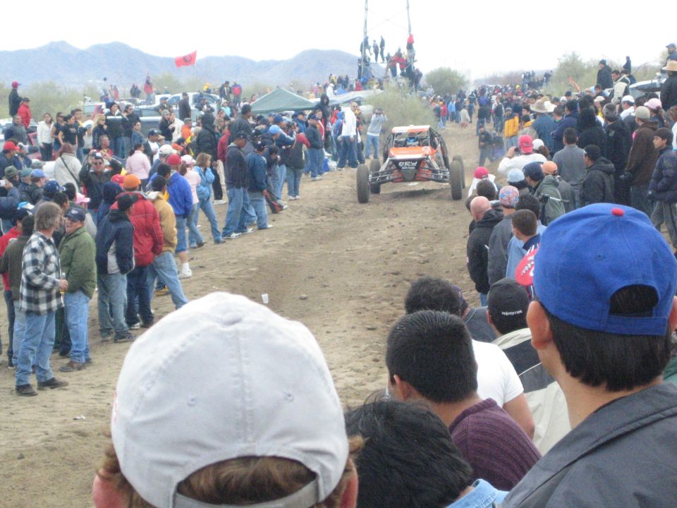 Off-road buggy on dirt course with crowds and red flags on a distant rise