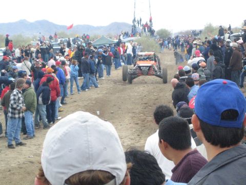 1990 Ford Bronco - off-road buggy lifts its front wheels on a dirt desert course as a thick crowd of spectators watches from both sides.
