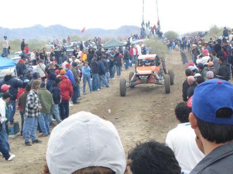 1990 Ford Bronco - orange and black off-road buggy marked 'OEW' races a dirt desert course bordered by rows of bundled-up spectators.
