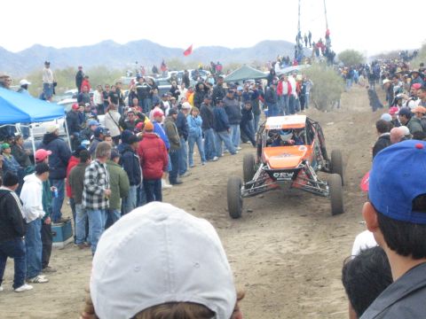 1990 Ford Bronco - orange off-road buggy lettered 'OEW' crests a dirt rise on a desert course as spectators look on from the sidelines.