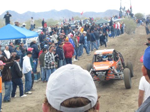 1990 Ford Bronco - orange and black off-road buggy drives a dirt desert course while a large crowd of spectators lines the track under a blue canopy.