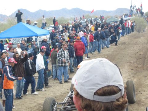1990 Ford Bronco - Crowds of spectators in jackets line a dirt desert race course near a blue canopy, with mountains under overcast skies.