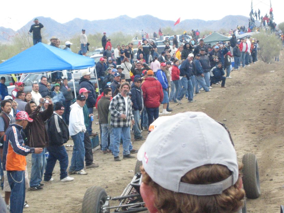 Spectators line a desert dirt course near a blue canopy under gray skies