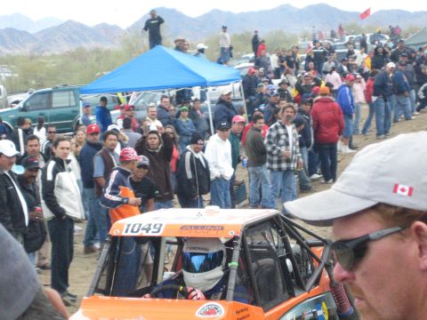 1990 Ford Bronco - orange off-road buggy numbered 1049 with a helmeted driver sits in a staging area as spectators crowd around it.