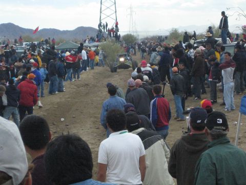 1990 Ford Bronco - off-road vehicle with its headlights on travels a dusty desert course as dense crowds of spectators watch from both sides.