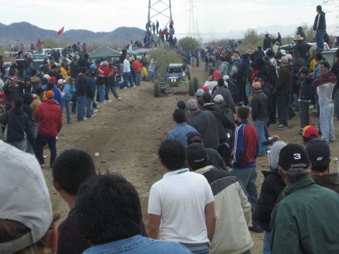 1990 Ford Bronco - off-road buggy runs a dirt desert course flanked by large crowds of spectators, with power-line towers and mountains behind.