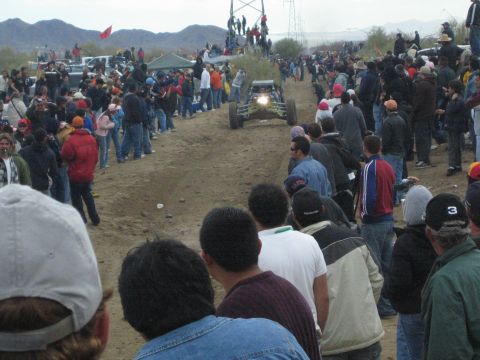 1990 Ford Bronco - off-road buggy drives a dirt desert course as crowds line the track and a red flag flies near power-line towers behind.
