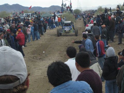 1990 Ford Bronco - blue tube-frame off-road buggy races down a dirt track flanked by a dense crowd of spectators with desert mountains behind.