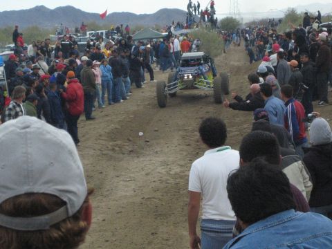 1990 Ford Bronco - blue single-seat off-road buggy heads down a sandy course as spectators line both sides under a cloudy desert sky.