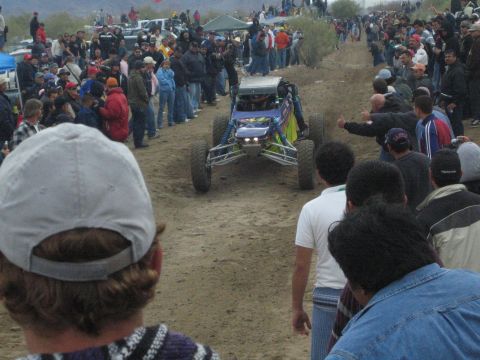 1990 Ford Bronco - blue off-road buggy with bright headlights drives down a dirt track surrounded by a large crowd of spectators.