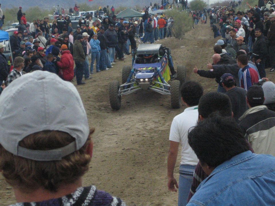 Blue off-road buggy with headlights on the dirt course among a crowd