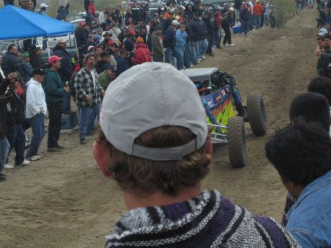 1990 Ford Bronco - buggy with a vivid multicolor paint scheme passes a crowd along a dirt course, seen over a spectator's gray backward cap.