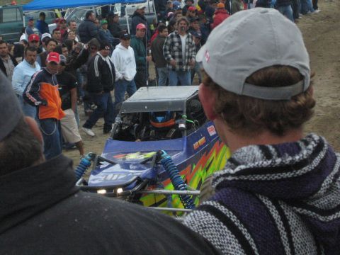 1990 Ford Bronco - close view of a blue and green off-road buggy with a helmeted driver, framed by spectators in the desert.