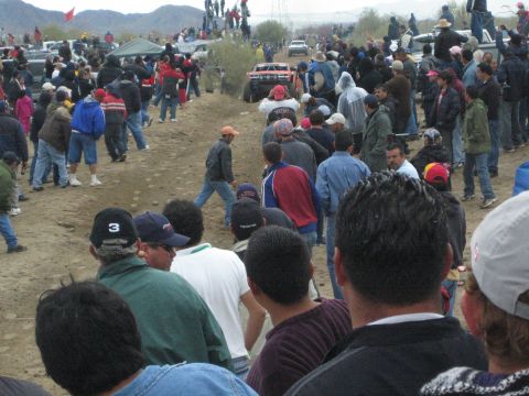 1990 Ford Bronco - red off-road race truck runs along a distant dirt course as spectators in caps watch from the foreground in the desert.