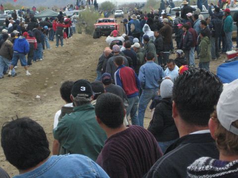 1990 Ford Bronco - red off-road truck travels a dirt course while a large crowd, including a cameraman, watches in the desert.