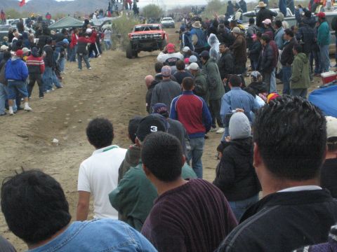 1990 Ford Bronco - red off-road race truck approaches down a sandy course flanked by spectators with desert mountains behind.