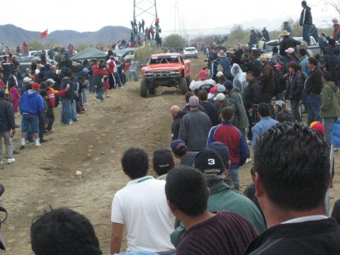 1990 Ford Bronco - red off-road race truck drives down a wide dirt course lined with spectators under a gray desert sky.