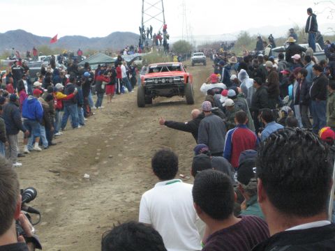 1990 Ford Bronco - orange-red off-road race truck heads down a dirt course as a crowd cheers and gives a thumbs-up in the desert.