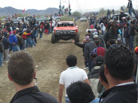 1990 Ford Bronco - Red off-road race truck number 800 drives down a dirt course lined with cheering spectators in the desert.