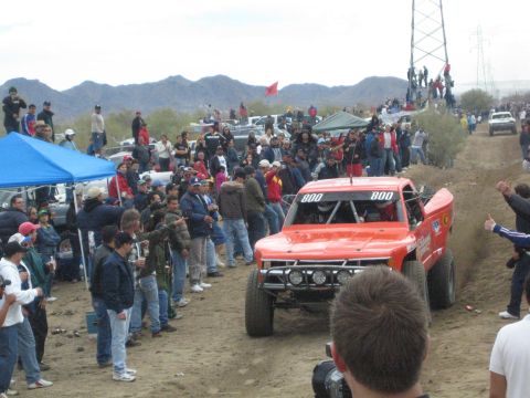 1990 Ford Bronco - Red off-road race truck number 800 faces the camera on a dirt course as spectators gather near a blue canopy tent.