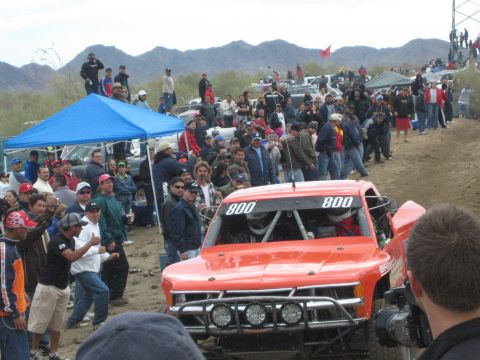 1990 Ford Bronco - front of red off-road race truck number 800 sits on a dirt course before a crowd and a blue canopy tent.