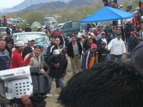 1990 Ford Bronco - crowd of spectators gathers in the desert near vehicles and a blue tent while a man films with a camcorder.