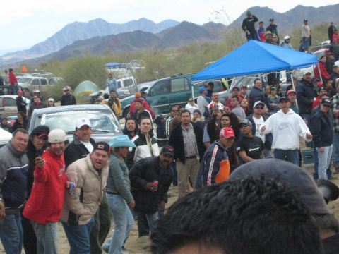1990 Ford Bronco - large crowd of spectators stands among parked trucks and a blue tent in the desert with mountains behind.