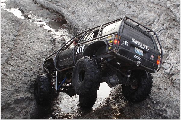 Black caged Jeep Cherokee XJ crawler on big mud tires climbing wet rocks, rear three-quarter view