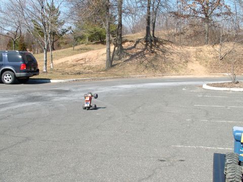 1994 Ford Bronco - A remote control car is seen on a paved surface next to a parked blue SUV, with trees and a grassy hill in the background.