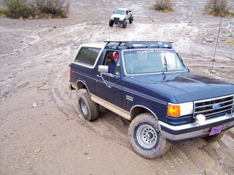 1991 Ford Bronco - A blue Ford Bronco navigates a muddy off-road course, with another white truck in the background.