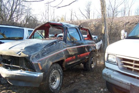 1977 Ford F-150 - This heavily damaged black pickup truck sits in a junkyard next to other vehicles.