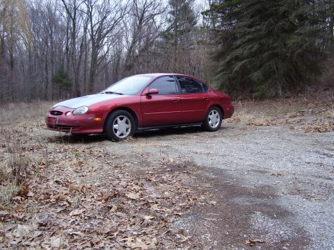 1997 Ford Taurus - A red sedan with a gray hood sits on a gravel lot surrounded by trees.