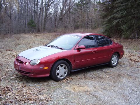 1997 Ford Taurus - A red sedan with a silver hood is parked outdoors in a wooded area. The vehicle appears to be a 1997 Ford Taurus.