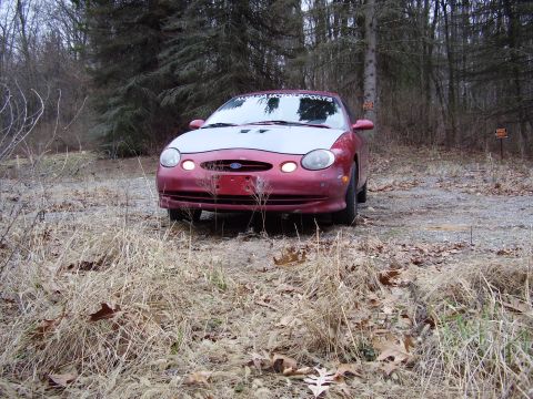 1997 Ford Taurus - A red Ford Taurus with a silver hood sits on a wooded path, its headlights on.