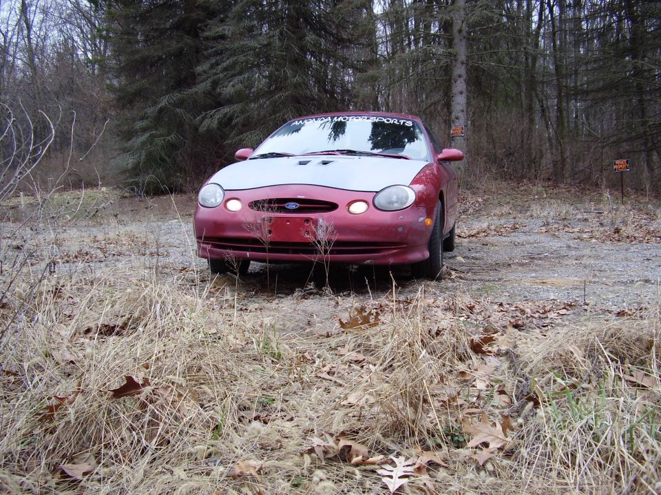 Red Ford Taurus with a silver hood parked on a dirt road in the woods