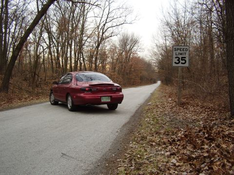 1997 Ford Taurus - A red 1997 Ford Taurus drives down a tree-lined road with a 35 mph speed limit sign.