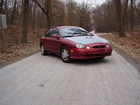 1997 Ford Taurus - A red sedan with a gray hood sits on a paved road surrounded by trees in the fall.
