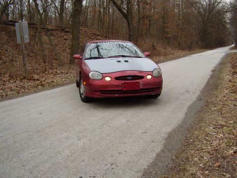 1997 Ford Taurus - A red 1997 Ford Taurus with a grey hood drives down a rural road surrounded by trees.