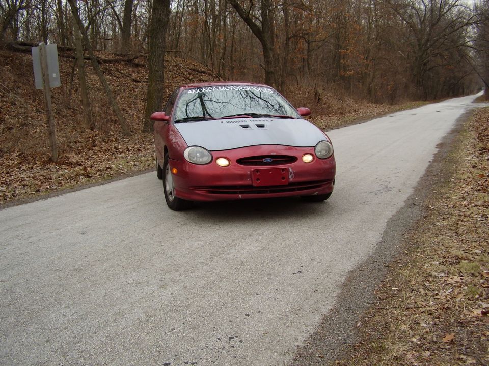 Red 1997 Ford Taurus with a grey hood and aftermarket hood scoops, driving on a paved road through a wooded area