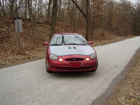 1997 Ford Taurus - A red 1997 Ford Taurus with a silver hood is parked on a rural road, with trees lining the background.