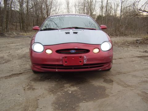 1997 Ford Taurus - A red Ford Taurus with a grey hood sits on a dirt road, facing forward with headlights on.