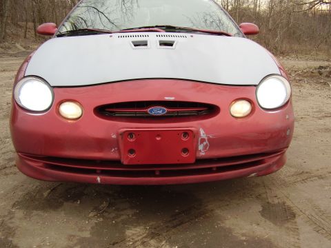 1997 Ford Taurus - Front view of a red third-generation Ford Taurus with raised hood, oval grille emblem and headlights on, parked on dirt.